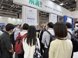 Crowded exhibition hall at a trade show with white booths and hanging signage in Japanese.