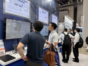 Trade show booth with multiple screens displaying schedules and charts; attendees observe the data while standing at the counter.