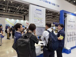 Crowded trade show booth with a large YASKAWA poster, white and blue display stands, and attendees in blue uniforms talking at the booth.