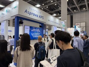 Busy trade-show booth for Asprova APS with large blue banner and screens, surrounded by attendees.