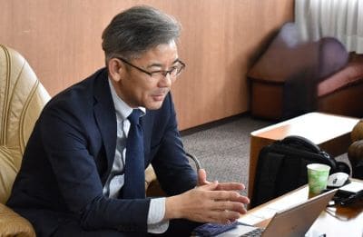 Businessman wearing glasses in a dark suit sits at a desk, gesturing with his hands while looking at a laptop in an office setting.