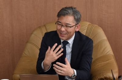 Man in a dark suit and light tie seated in a tan leather armchair, gesturing with his hands during a discussion.