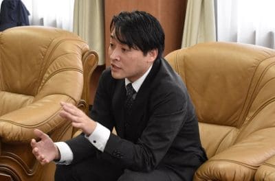 Man in a dark suit sits on a tan leather couch, gesturing with open hands during a discussion in an office setting.