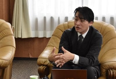 Man in a dark suit sits in a brown leather chair, talking and gesturing with his hands during a meeting in an office setting with a laptop nearby.