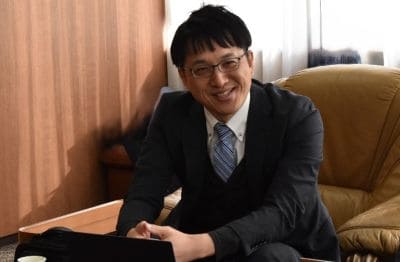 Smiling man in a dark suit and blue tie sits on a brown leather sofa, working on a laptop on a nearby table.