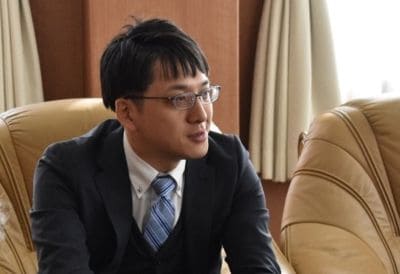 Man wearing glasses and a dark suit sits in a brown leather chair in an office, looking to the side as if speaking.