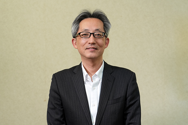 Portrait of a smiling Asian man in a dark pinstripe suit and white shirt, standing against a beige wall.