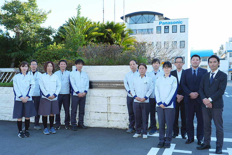 Twelve professionals in white jackets and business attire stand in a parking lot outside a Panasonic building with greenery.