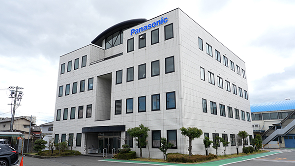 Panasonic corporate building with a white stone exterior, blue Panasonic sign, three stories, glass entrance, and surrounding trees in a parking lot.