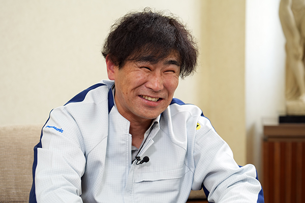 Smiling man in a light blue work shirt sits indoors, wearing a small microphone on his chest.