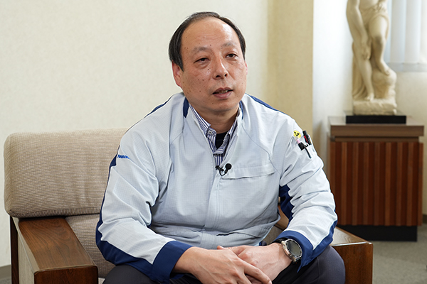 Middle-aged man in a light blue work shirt sits on a wooden-armchair, speaking with a lapel mic in an office setting.