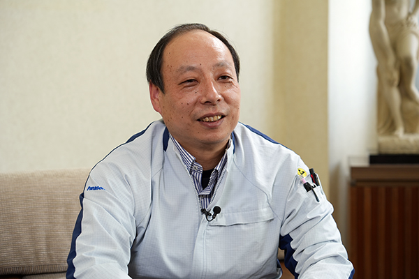 Smiling man in a light blue work jacket seated on a sofa indoors, speaking into a lapel microphone during an interview.