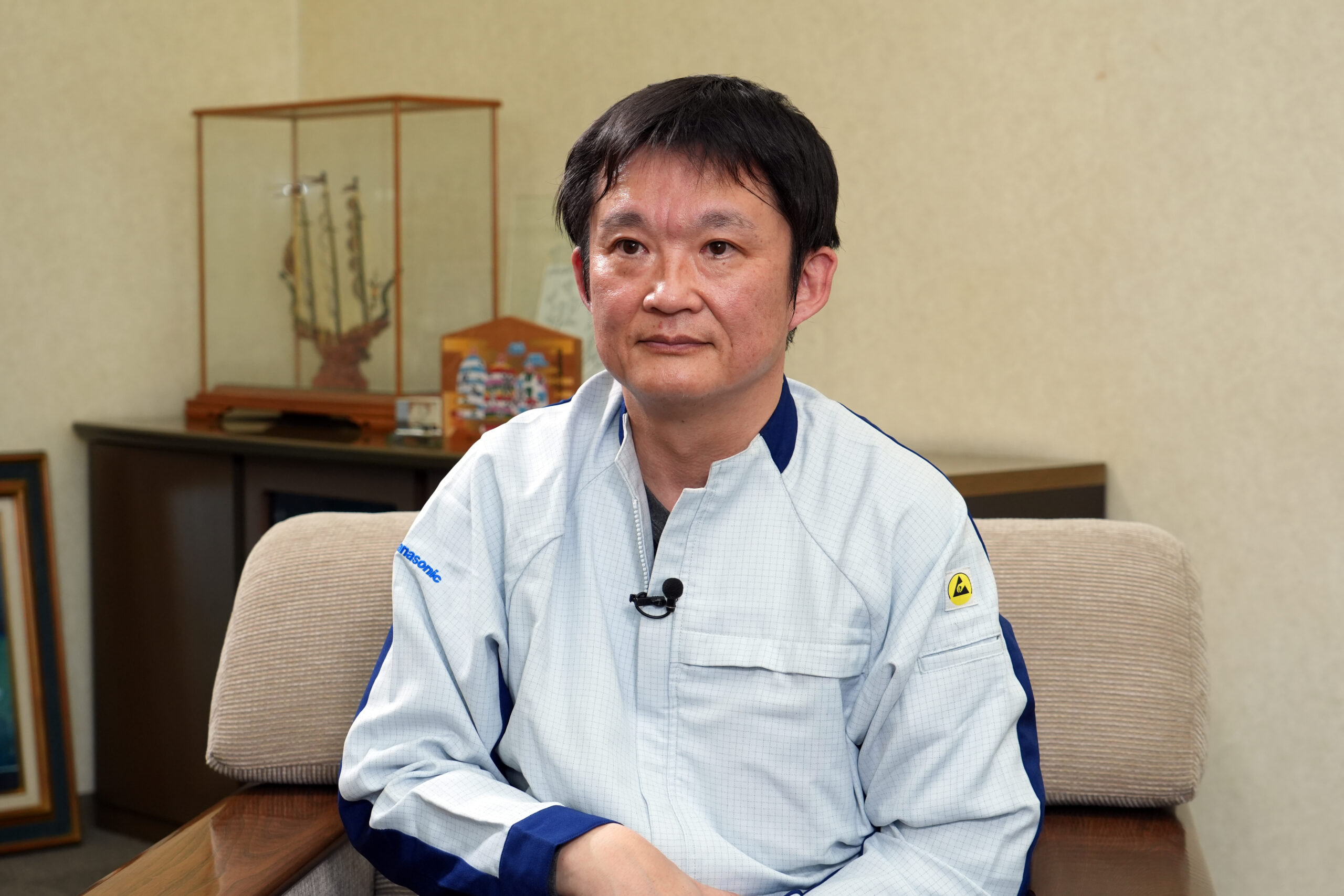 Man wearing a light blue work shirt sits on a beige sofa, hands folded, during an interview setting with a microphone clipped to his chest.