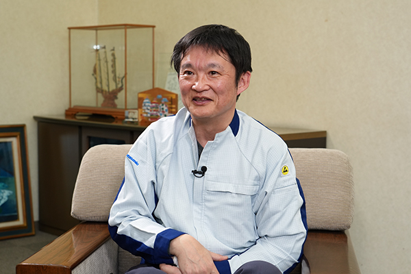 Asian man in a light blue work shirt sits on a beige sofa in an office, with a clip-on microphone attached to his shirt.