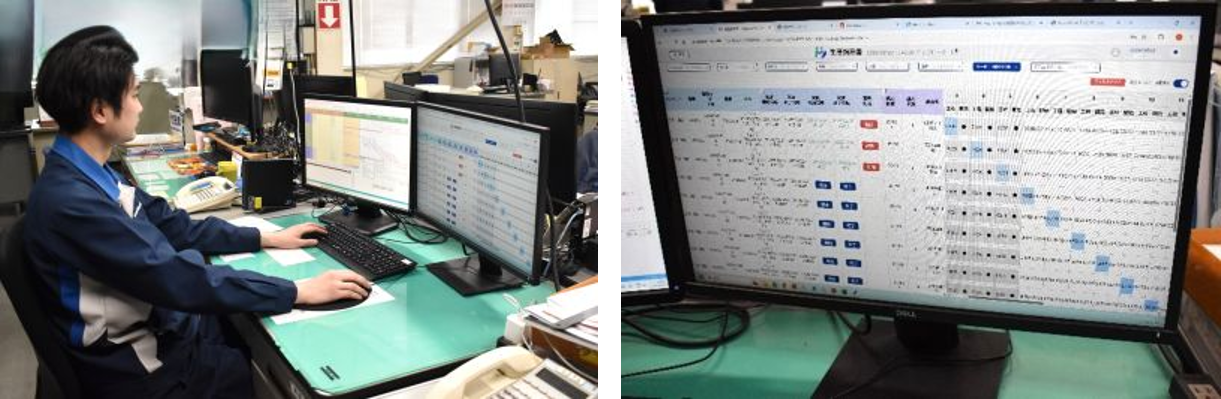 A worker sits at a desk with two computer monitors, typing on a keyboard in a workroom; the left screen shows a spreadsheet or dashboard. The right screen is a close-up of a monitor displaying a dense data table or list of entries.
