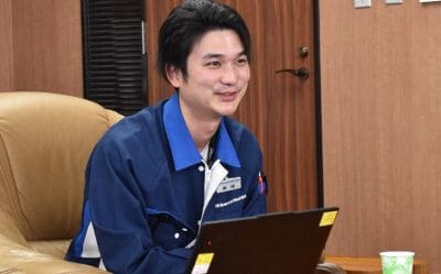 Young man in a blue work uniform sitting on a sofa with a laptop, smiling in an office setting.