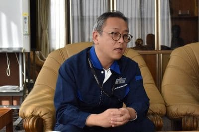 Man in a blue shirt with glasses sits on a tan leather sofa, speaking in an office-like room.