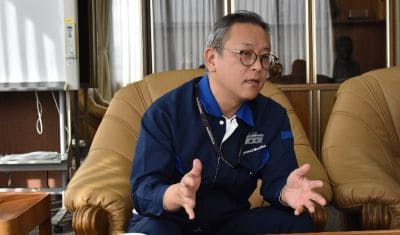 Man wearing a blue shirt and glasses sits on a tan leather couch, gesturing with his hands as he speaks during a discussion.