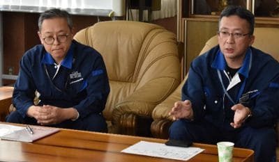 Two men in blue jackets sit at a wooden table, discussing documents in an office setting.