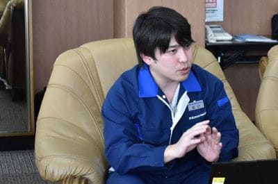 A man in a blue uniform sits in a tan leather chair in an office, gesturing with his hands while looking to the side.