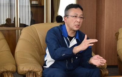 Man in a blue jacket sits on a tan leather chair in an office, speaking and gesturing with his hands during a discussion.
