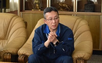 Asian man wearing glasses in a blue jacket sits on a beige leather sofa, hands clasped, speaking in a lounge/office setting.