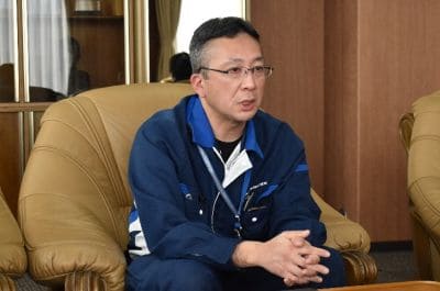 Man in a blue jacket sits on a beige armchair, hands clasped, speaking in a meeting room.