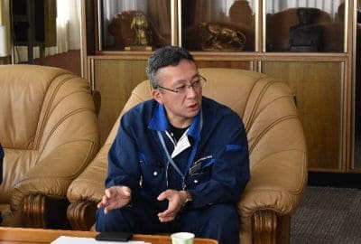 Man in a blue jacket seated on a tan leather sofa, gesturing with his hands while speaking in an office setting with display cabinets behind him.