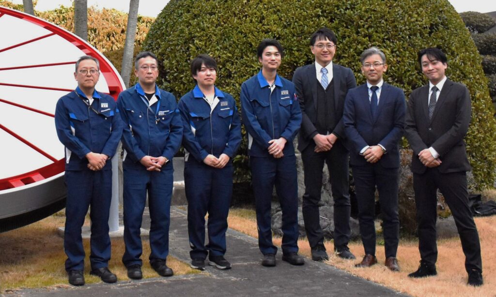 Eight men in blue work uniforms and dark suits pose outdoors beside a large red circular sculpture and green hedges.