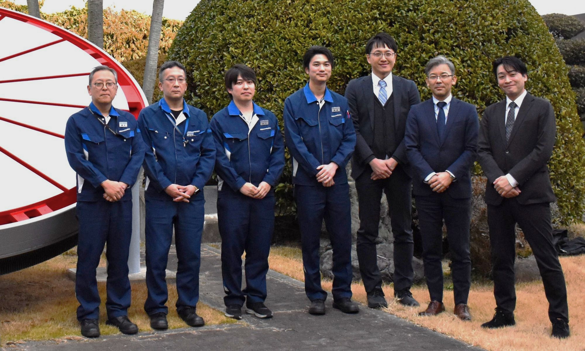 Eight men in blue work uniforms and dark suits pose outdoors beside a large red circular sculpture and green hedges.