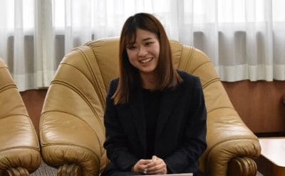 Smiling woman in a dark blazer sits in a large beige armchair during a formal interview or meeting.